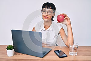 Beautiful brunettte woman working at the office eating healthy apple thinking attitude and sober expression looking self confident