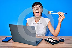 Beautiful brunettte woman working at the office eating sushi angry and mad screaming frustrated and furious, shouting with anger