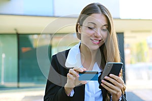 Beautiful business woman holds modern cell phone and credit card making shopping online outdoor office on break time. People and