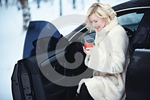 Beautiful business woman in luxurious white fur coat drinking hot coffee on snowy winter day sitting in her car