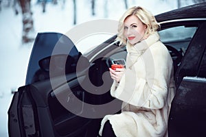 Beautiful business woman in luxurious white fur coat drinking hot coffee on snowy winter day sitting in her car