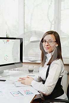 Beautiful business woman in suit and glasses working at computer with documents in light office