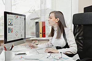 Beautiful business woman in suit and glasses working at computer with documents in light office