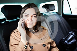 Beautiful business woman is using a smart phone and smiling while sitting on back seat in the car