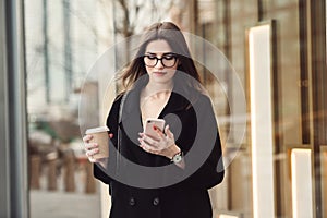 Beautiful business woman wearing glasses walking to the work with coffee and texting on phone outdoors.