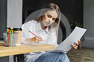 Beautiful business woman working on project while sitting at wooden table, makes notes on white sheet of paper.