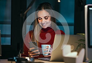 Beautiful business young woman using her mobile phone while working with laptop in the office at night