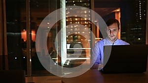 Beautiful businessman working overtime at night in executive office. City lights are visible in background from a large