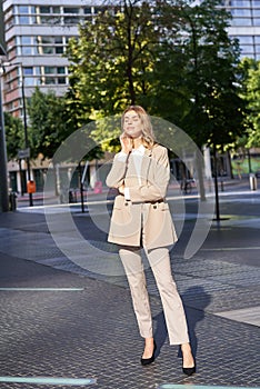 Beautiful businesswoman in beige suit, standing near office buildings on street, posing with confident, relaxed face