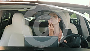 Beautiful businesswoman looking in rear view mirror and making up her lips with lipstick in car. Transport, trip