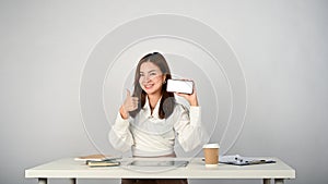 A beautiful businesswoman sits at her desk, showing her thumb up and holding a smartphone