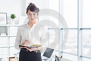 Beautiful businesswoman standing in office, holding notebook, planning meetings for the work day, looking at camera.