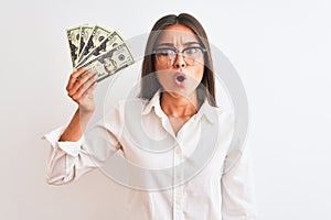 Beautiful businesswoman wearing glasses holding dollars over isolated white background scared in shock with a surprise face,