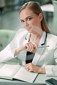 Beautiful businesswoman in white jacket looking at camera making notes in her scheduler or journal sitting in front at