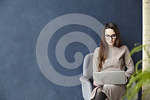 Beautiful businesswoman working on laptop while sitting in modern loft office. Dark blue wall background, day light.