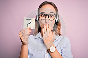 Beautiful call center agent woman working using headset holding reminder with question mark cover mouth with hand shocked with