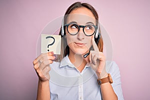 Beautiful call center agent woman working using headset holding reminder with question mark serious face thinking about question,