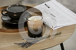Beautiful candle, lavender, newspaper and cup of coffee on round wooden table indoors
