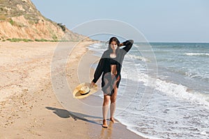 Beautiful carefree woman with hat walking on sandy beach at sea waves and relaxing. Summer vacation. Fit stylish young female in