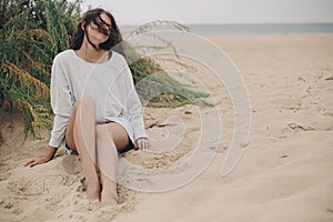 Beautiful carefree woman with windy hair and in sweater sitting on sandy beach on background of green grass and sea, tranquil