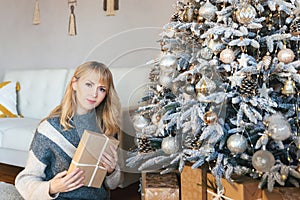 A beautiful Caucasian 40 years old woman with a gift in hands posing at Christmas tree. Classic christmas New Year