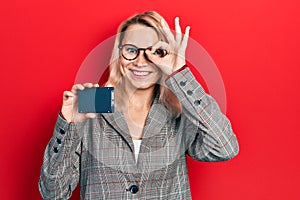 Beautiful caucasian blonde business woman holding ssd memory smiling happy doing ok sign with hand on eye looking through fingers