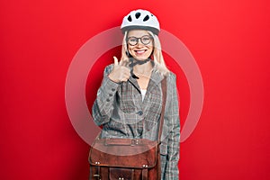 Beautiful caucasian blonde business woman wearing bike helmet doing happy thumbs up gesture with hand