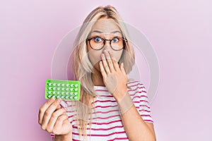 Beautiful caucasian blonde girl holding birth control pills covering mouth with hand, shocked and afraid for mistake