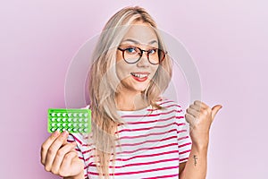 Beautiful caucasian blonde girl holding birth control pills pointing thumb up to the side smiling happy with open mouth