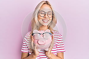 Beautiful caucasian blonde girl holding piggy bank with glasses smiling and laughing hard out loud because funny crazy joke