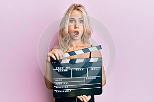Beautiful caucasian blonde girl holding video film clapboard afraid and shocked with surprise and amazed expression, fear and