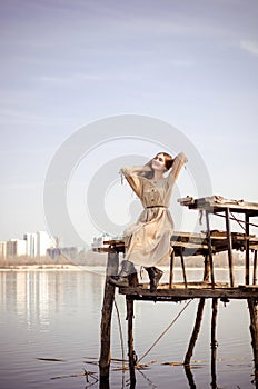 Beautiful caucasian blonde girl in light dress, sits on old wooden bridge over water against blue sky on city background