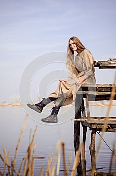 Beautiful caucasian blonde girl, in light dress, sits on old wooden bridge over water on background of blue sky