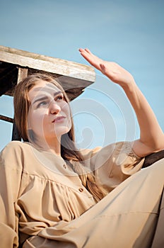 Beautiful caucasian blonde girl, in light dress, sits on old wooden bridge over water on background of blue sky