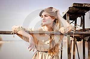 Beautiful caucasian blonde girl, in light dress, sits on old wooden bridge over water on background of blue sky