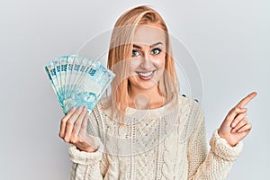 Beautiful caucasian blonde woman holding 100 brazilian real banknotes smiling happy pointing with hand and finger to the side