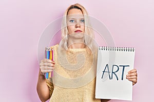 Beautiful caucasian blonde woman holding art notebook and color pencils relaxed with serious expression on face