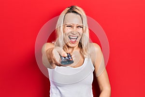 Beautiful caucasian blonde woman holding television remote control smiling and laughing hard out loud because funny crazy joke