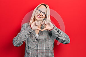 Beautiful caucasian blonde woman wearing business jacket and glasses smiling in love doing heart symbol shape with hands