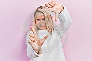 Beautiful caucasian blonde woman wearing casual winter sweater smiling making frame with hands and fingers with happy face