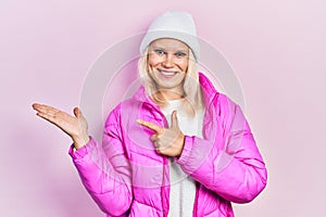 Beautiful caucasian blonde woman wearing wool hat and winter coat amazed and smiling to the camera while presenting with hand and