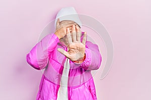 Beautiful caucasian blonde woman wearing wool hat and winter coat covering eyes with hands and doing stop gesture with sad and