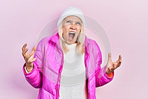 Beautiful caucasian blonde woman wearing wool hat and winter coat crazy and mad shouting and yelling with aggressive expression