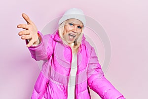 Beautiful caucasian blonde woman wearing wool hat and winter coat looking at the camera smiling with open arms for hug