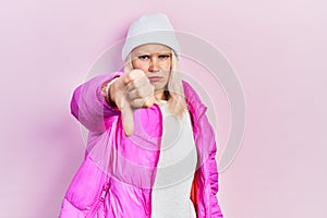 Beautiful caucasian blonde woman wearing wool hat and winter coat looking unhappy and angry showing rejection and negative with