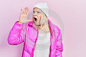 Beautiful caucasian blonde woman wearing wool hat and winter coat shouting and screaming loud to side with hand on mouth