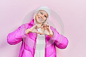 Beautiful caucasian blonde woman wearing wool hat and winter coat smiling in love doing heart symbol shape with hands