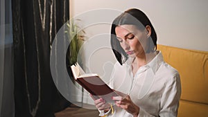 Beautiful Caucasian businesswoman, student, is studying at home, sitting on sofa and reading interesting book.