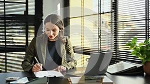 Beautiful caucasian female entrepreneur using laptop computer and analyzing financial document at her workplace