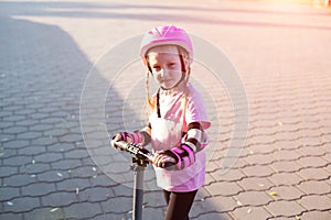 A beautiful caucasian girl of 7 years old in a pink helmet and armlets stands on a scooter. Summer sunny day.